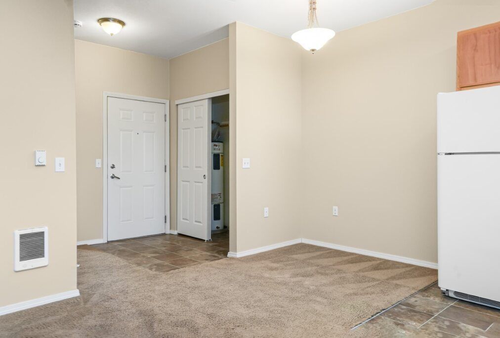 Empty beige apartment room with carpet, white doors, and a refrigerator at right.