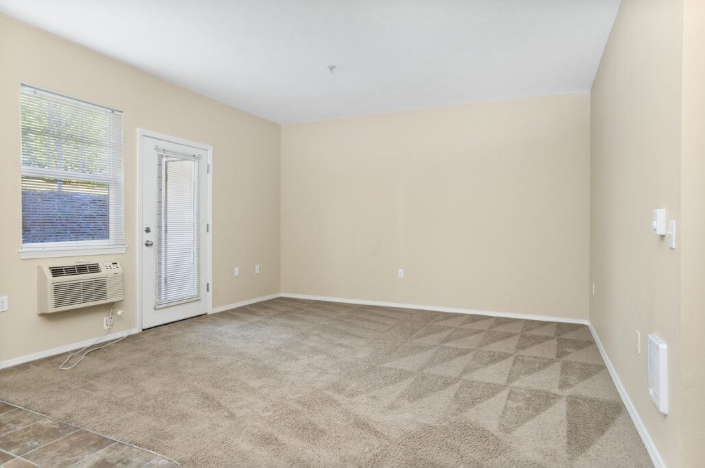 Empty beige carpeted room with cream walls, window, and wall-mounted air conditioner