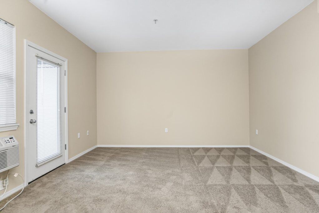 Empty beige living room with carpet, white trim, and a glass door near a window blind