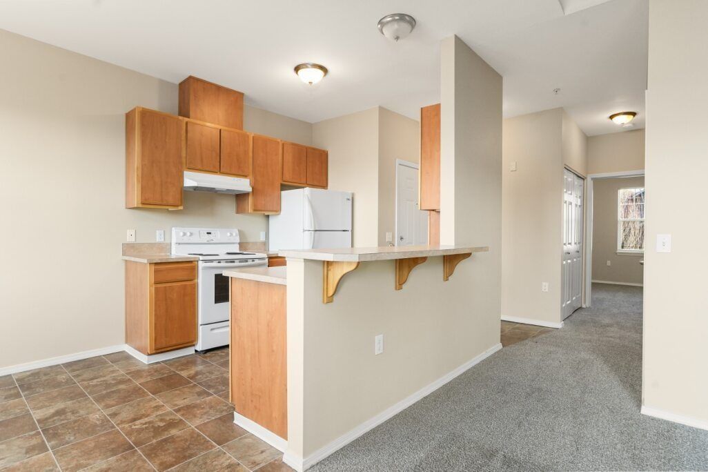 Empty apartment kitchen with wood cabinets, white appliances, and a carpeted hallway.