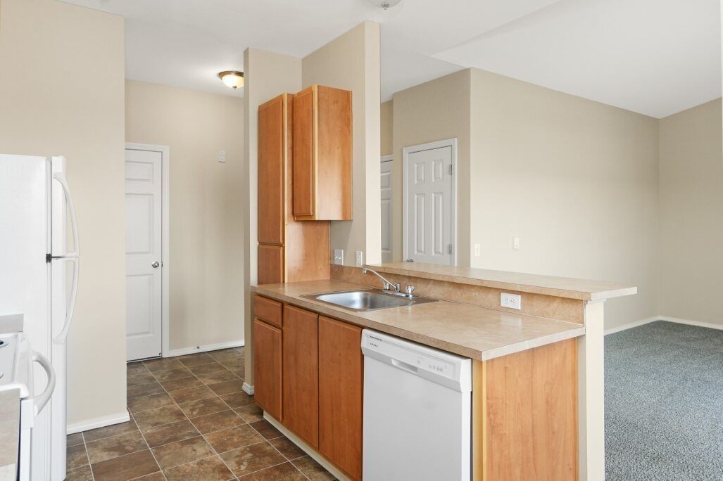 Empty apartment kitchen with wood cabinets, white appliances, and beige walls next to a carpeted living area