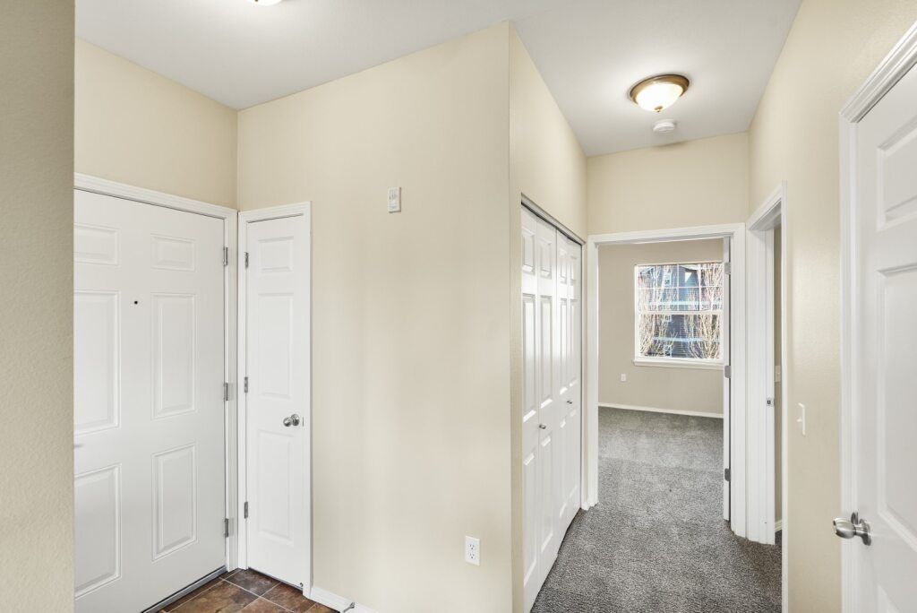 Bright hallway with white doors, beige walls, carpet, and a glimpse of a bedroom at the end.