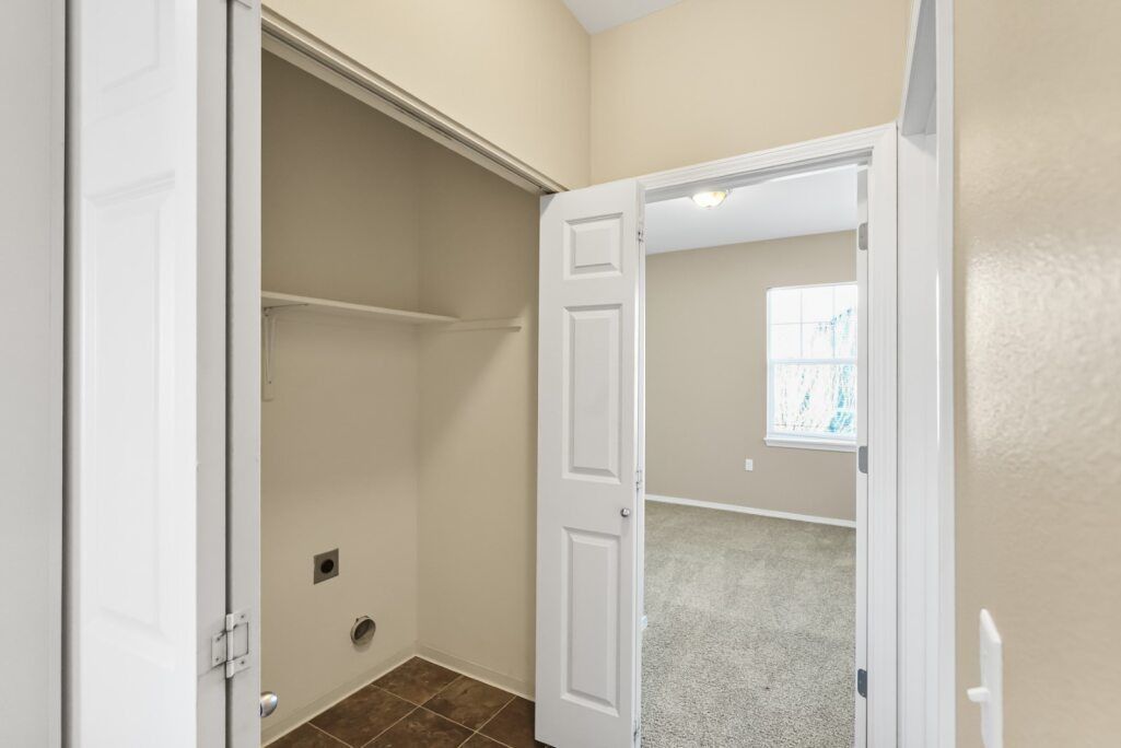 Empty closet with beige walls, tiled floor, and open bifold doors leading to a carpeted room.