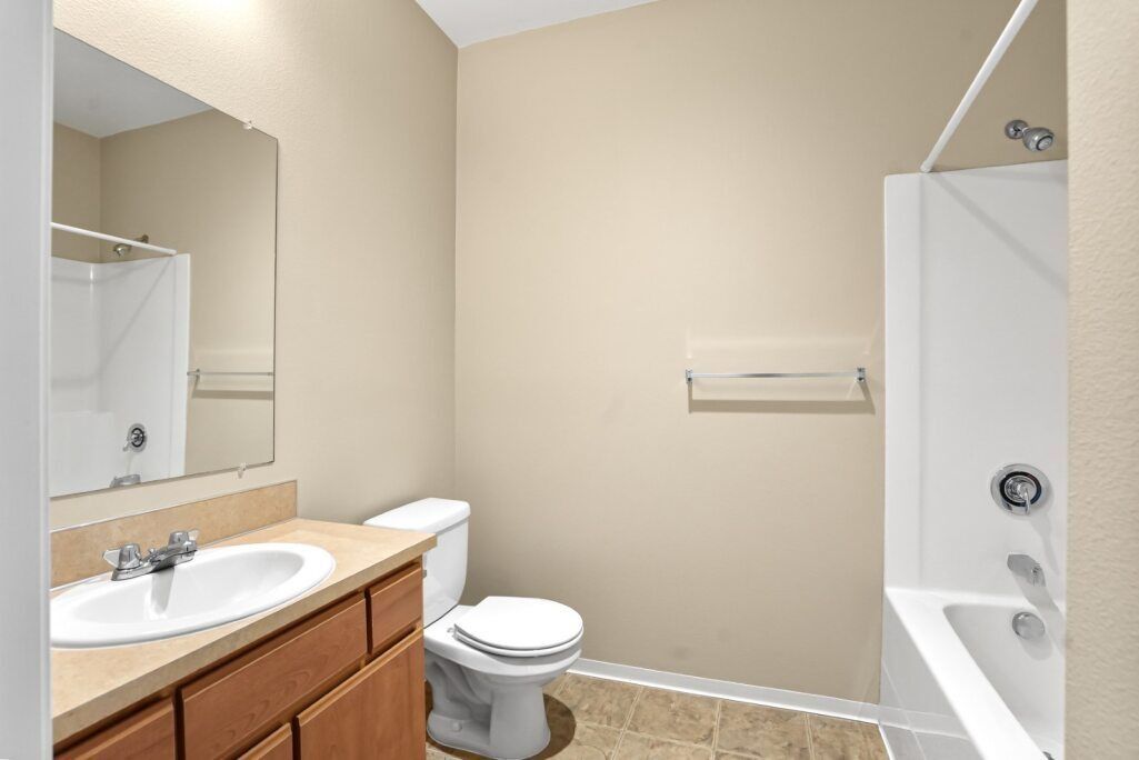Small beige bathroom with sink vanity, toilet, tub, and wall mirror.