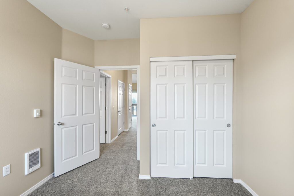 Empty beige bedroom with carpet, open door, mirrored closet, and hallway visible through doorway