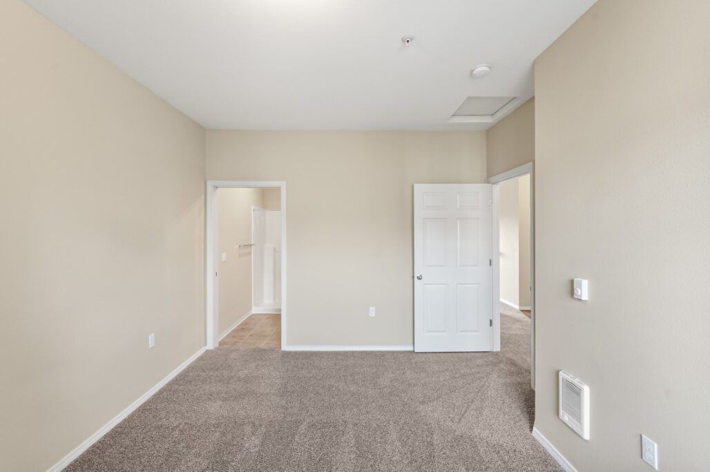 Empty beige bedroom with carpet, white closet doors, and open doorway to a hall
