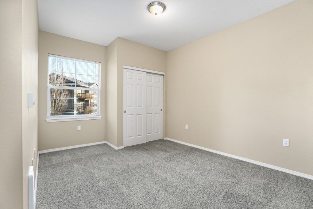 Empty beige bedroom with gray carpet, window, and white closet doors