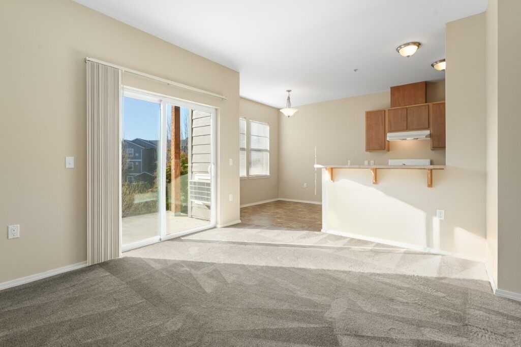 Empty carpeted living room with sliding glass door and a small kitchen counter in the corner