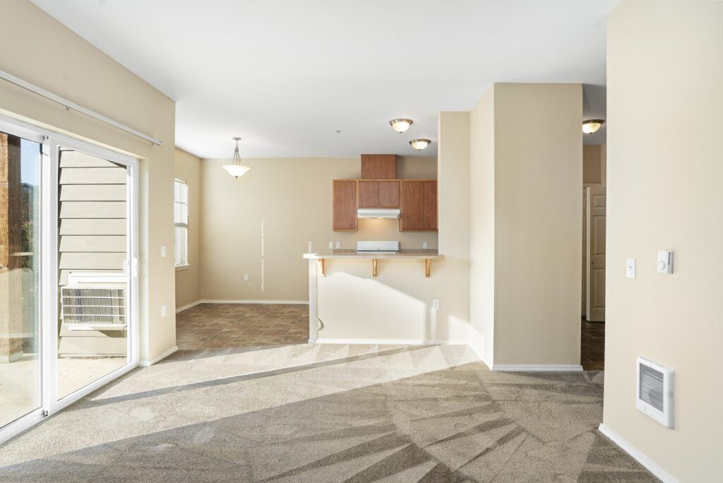Empty apartment living room with beige carpet, sliding glass doors, and a small kitchen with wood cabinets.