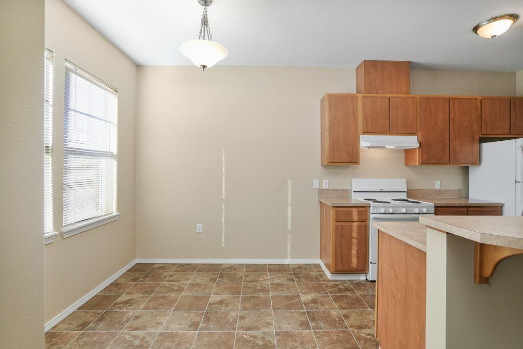 Empty kitchen with tan tile floor, white walls, and wood cabinets under a ceiling light.