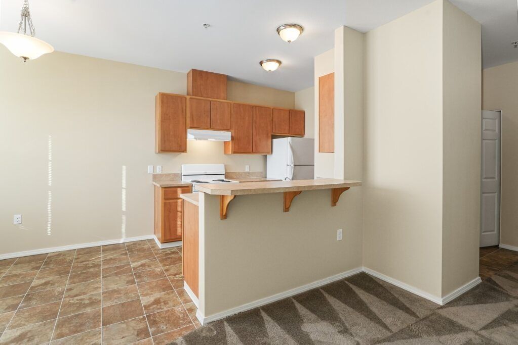 Empty kitchen with wooden cabinets, white appliances, and a peninsula counter in a tiled room.