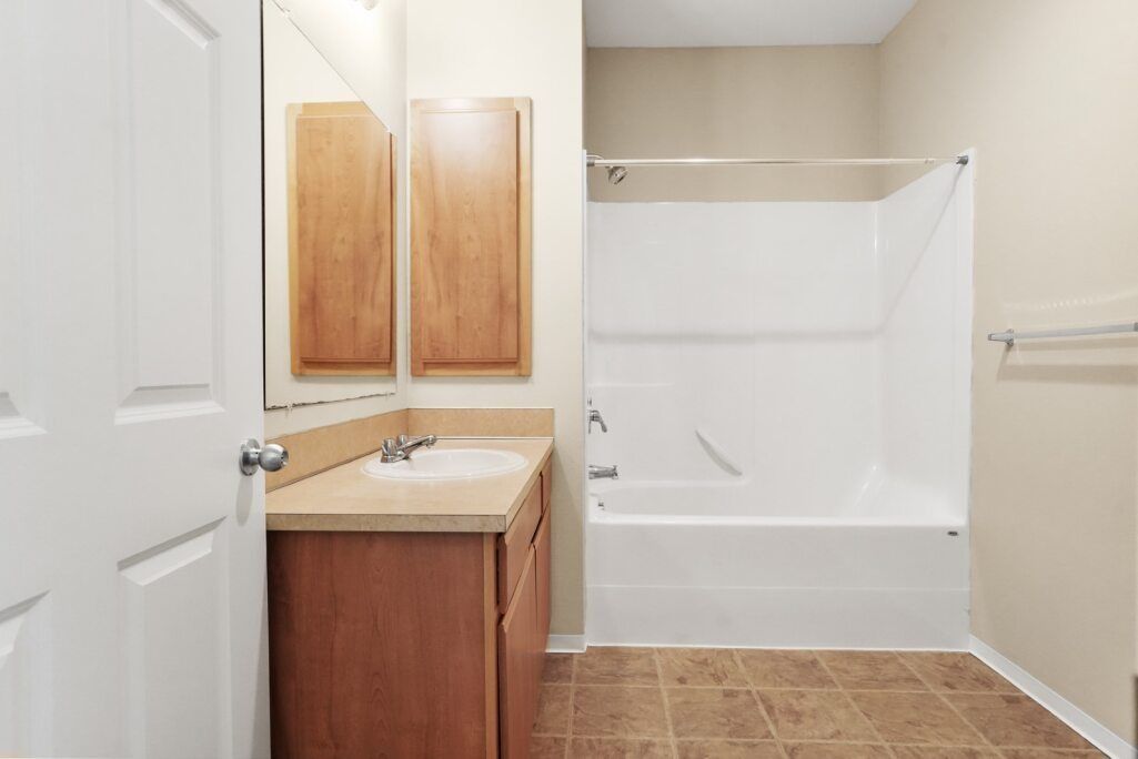 Small bathroom with tan tile floor, vanity, mirror, and white tub-shower combo.
