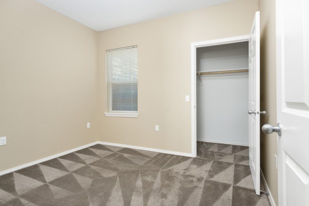 Empty beige bedroom with patterned carpet, window blinds, and open closet door