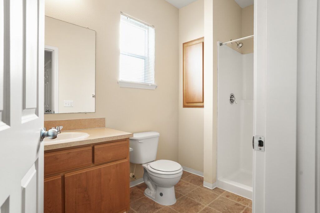Small beige bathroom with wood vanity, sink, toilet, window, and shower stall.