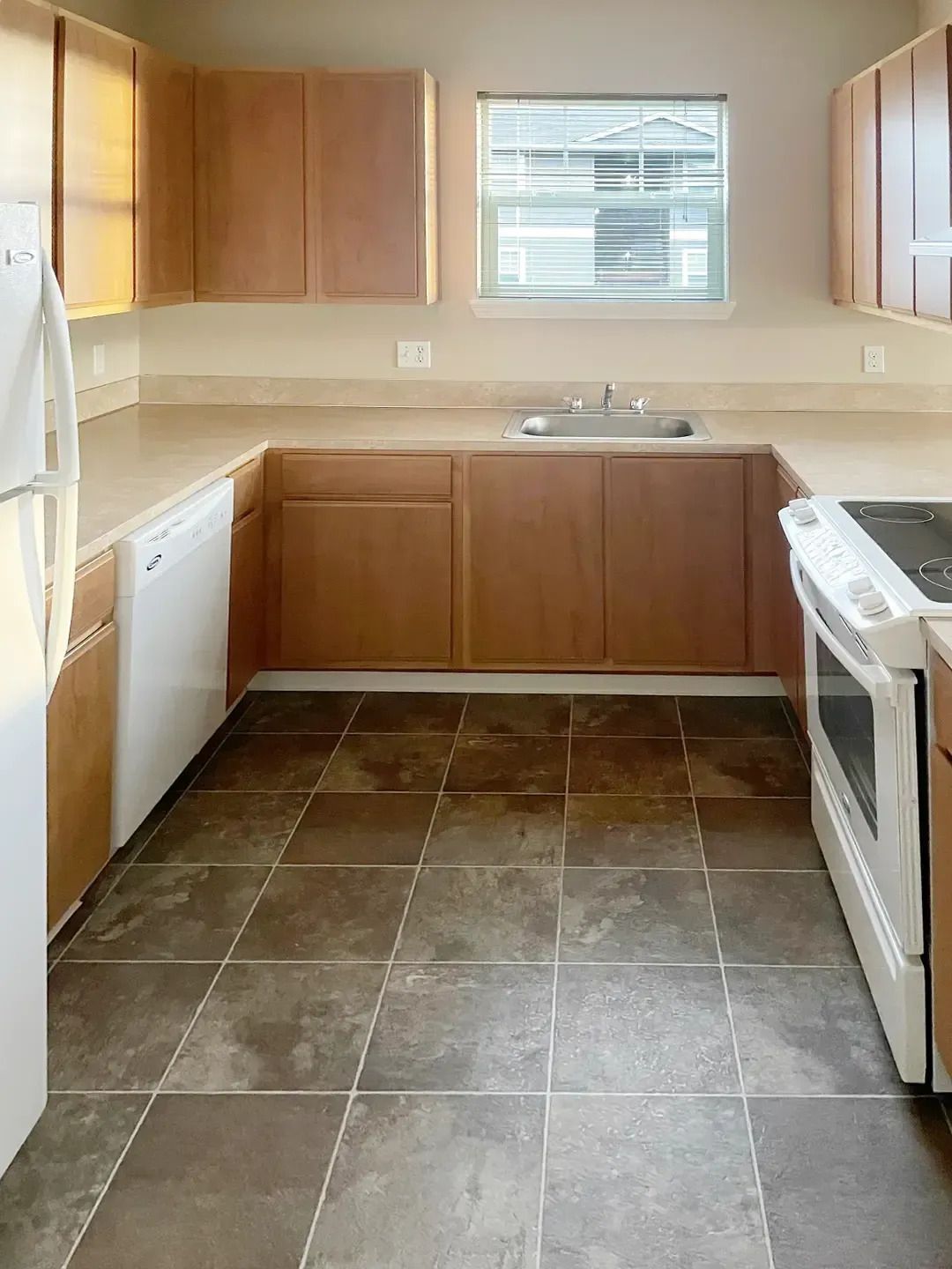Empty kitchen with wooden cabinets, white appliances, tiled floor, and a window above the sink.