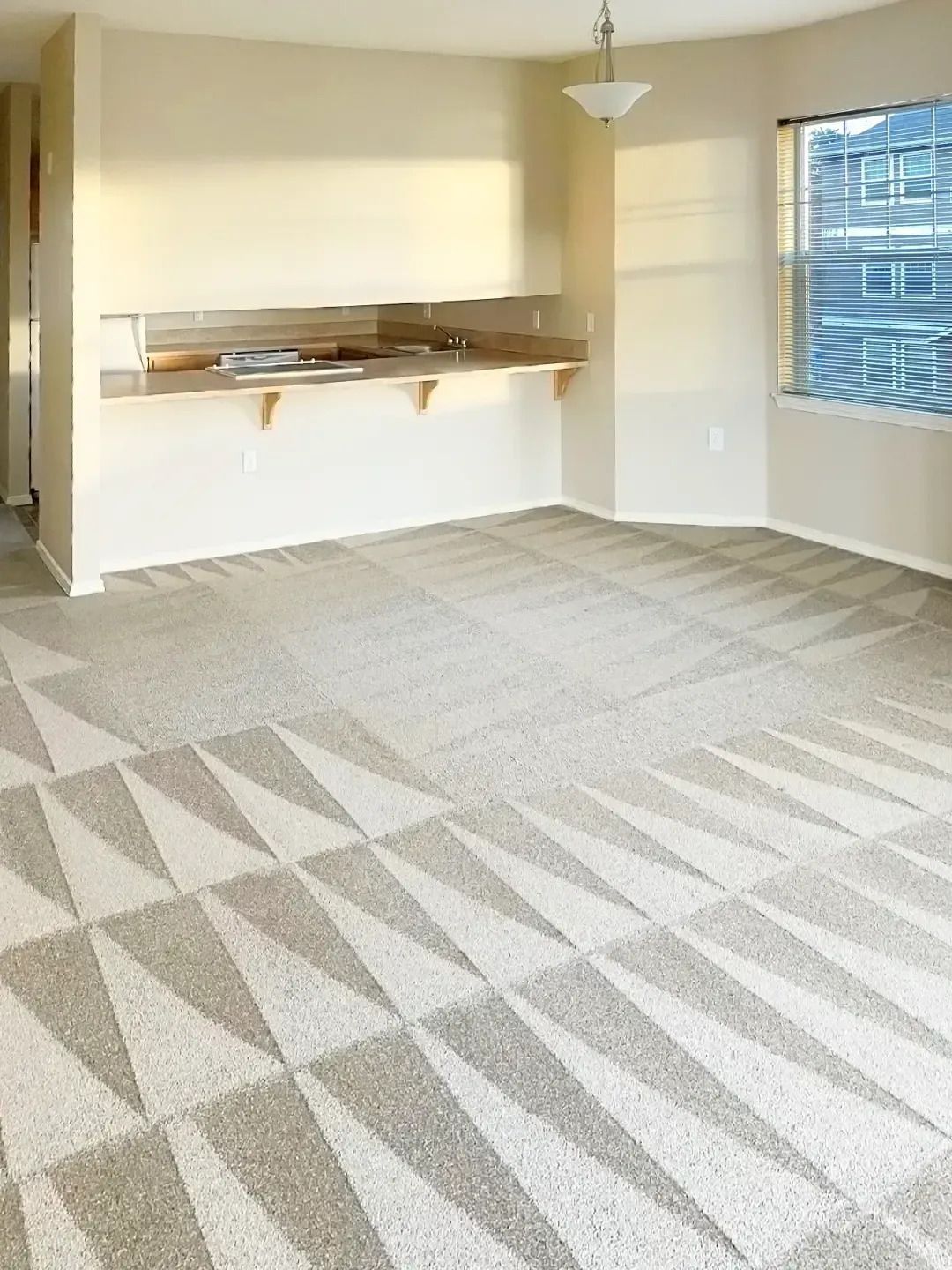 Empty beige living room with patterned carpet, wall shelf, and windows with blinds