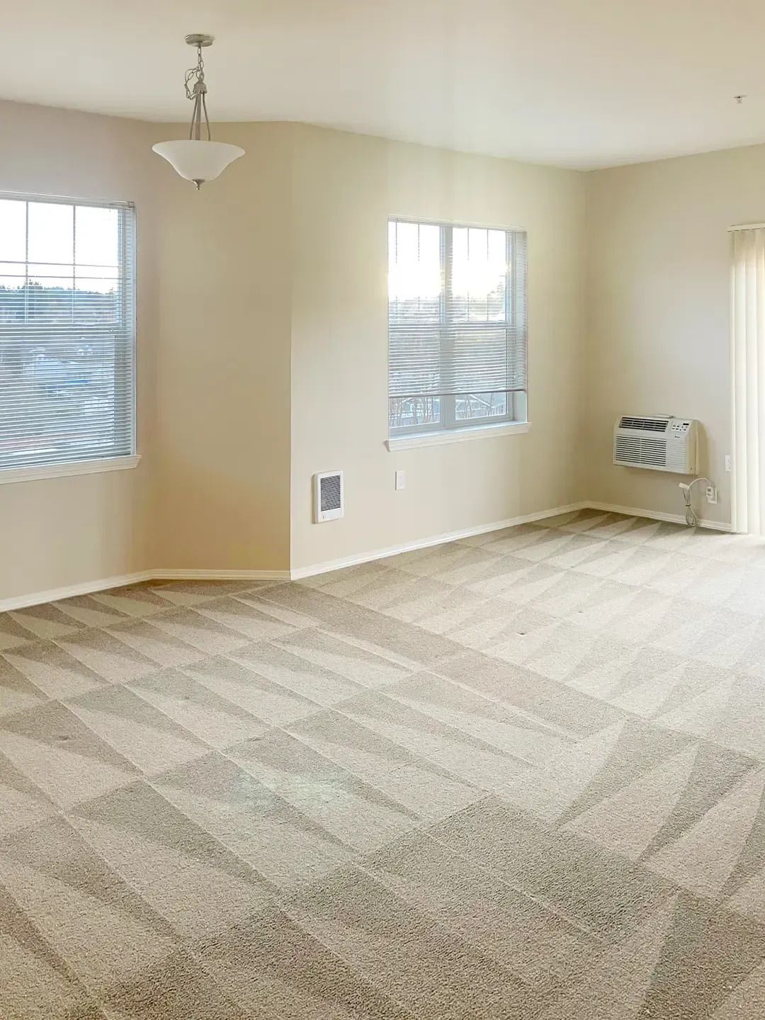 Empty beige living room with carpet, three windows, ceiling light, and wall-mounted air conditioner