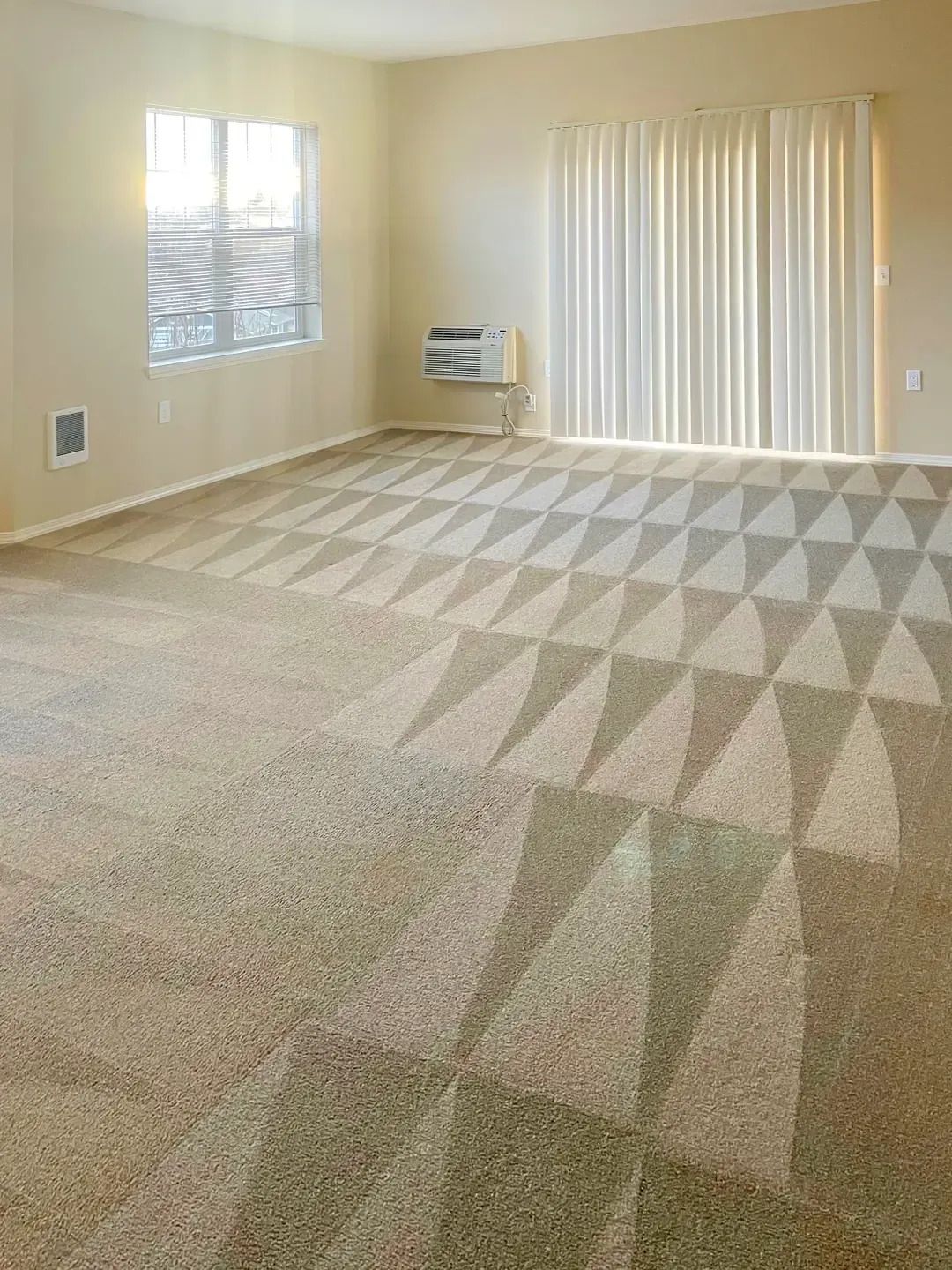 Empty beige carpeted room with a window, vertical blinds, and sunlight casting geometric shadows on the floor