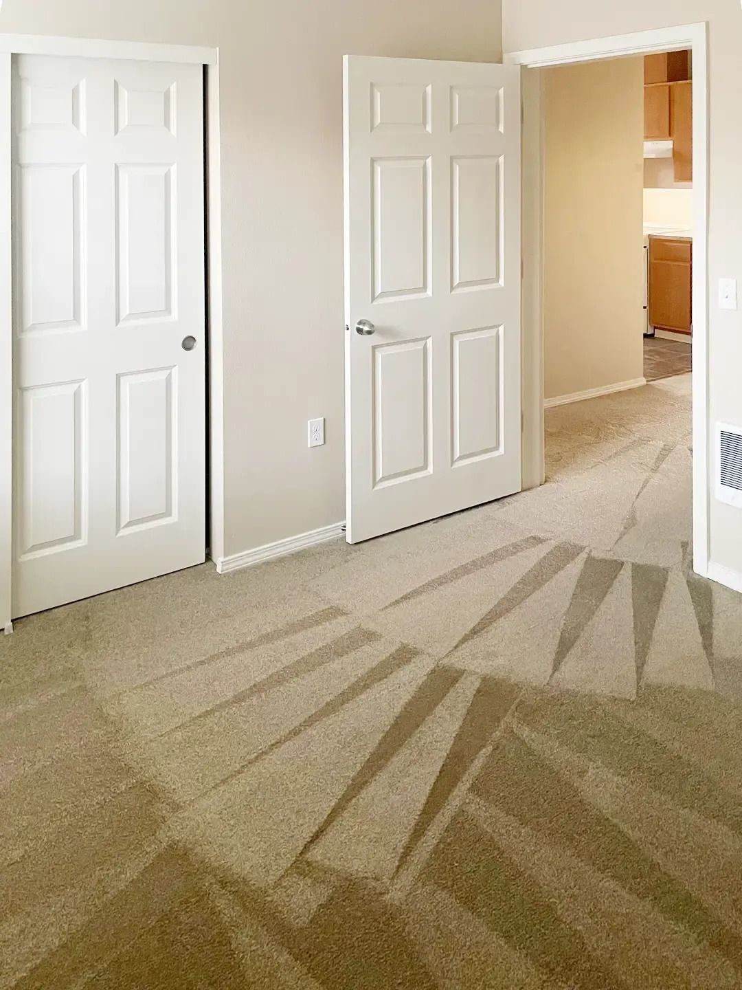Empty beige bedroom with two white doors and sunlight casting shadows on carpet.