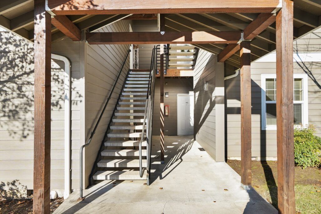 Covered outdoor staircase beside a building, with wooden posts and railings in bright sunlight