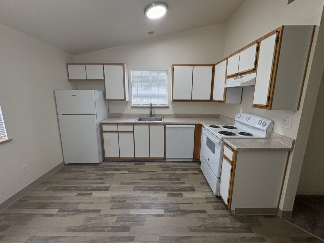 Empty kitchen with white cabinets, gray wood-look floors, and a stove, fridge, and sink under a window.