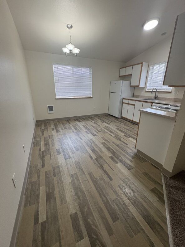 Empty kitchen with wood-pattern flooring, white cabinets, refrigerator, and ceiling light fixtures.