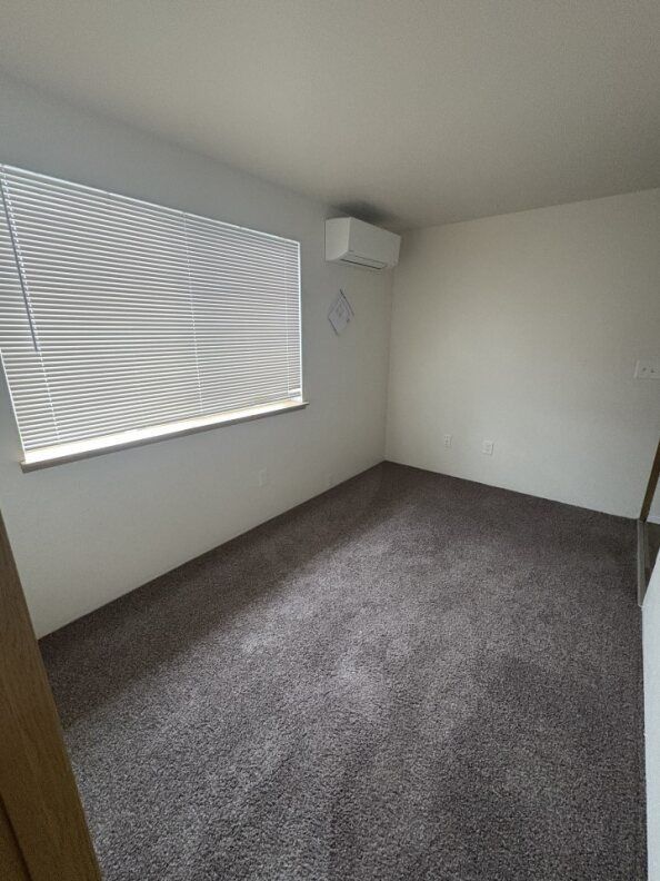 Empty carpeted room with a window and wall-mounted air conditioner.