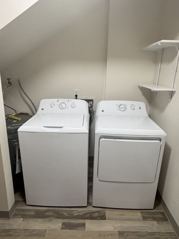 Laundry room with a white top-load washer and front-load dryer side by side, under a slanted wall.