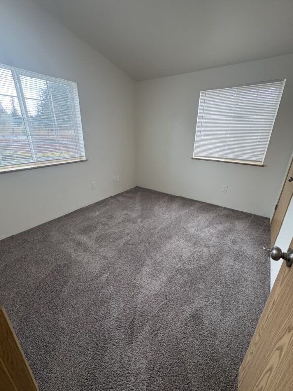 Empty carpeted room with white walls, two windows with blinds, and an open wooden door