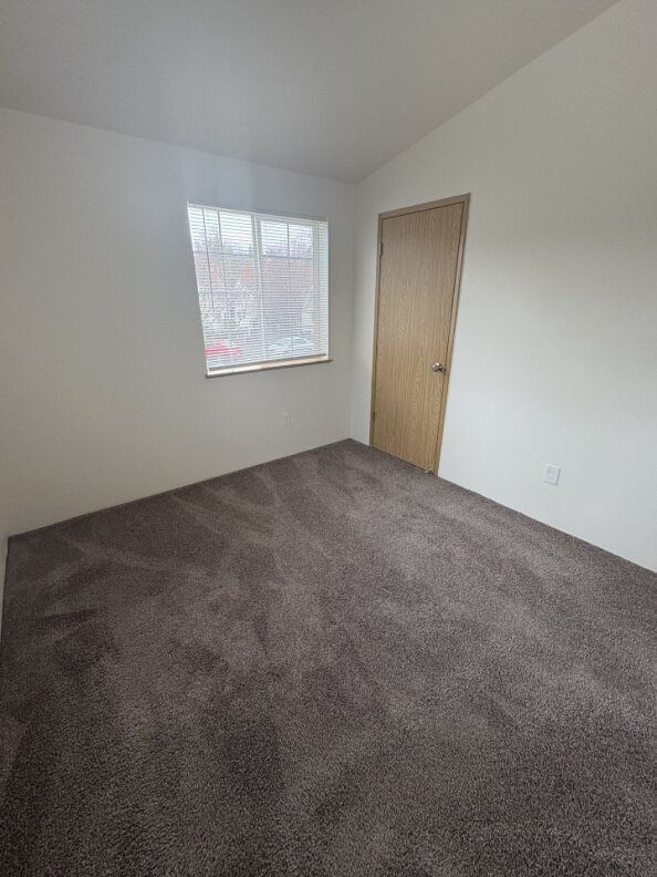 Empty carpeted room with white walls, a window, and a closed wooden door.