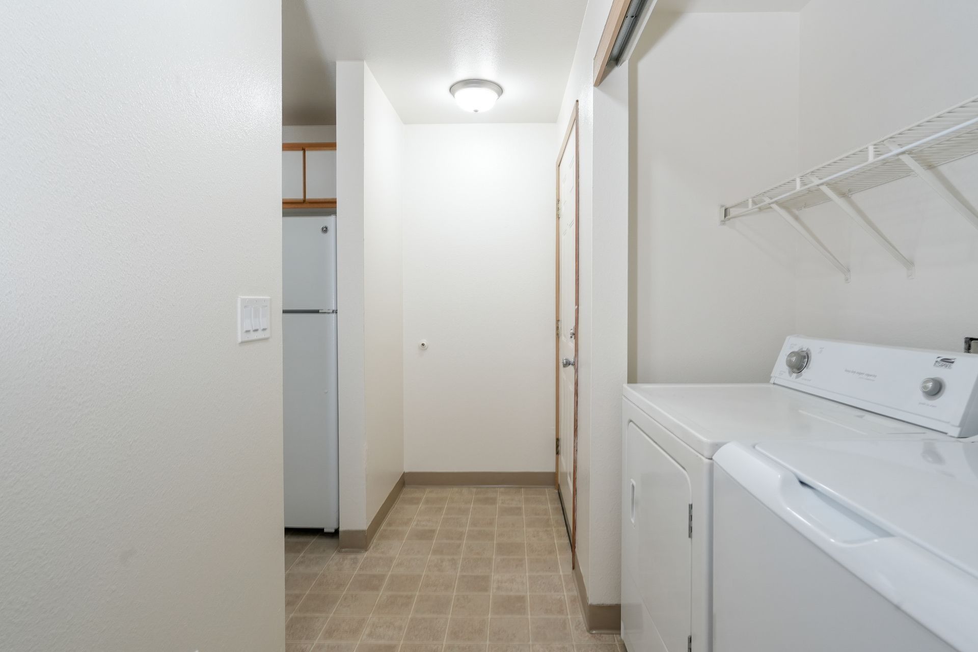 Narrow laundry hallway with washer and dryer, white walls, beige tile floor, and closet shelving.