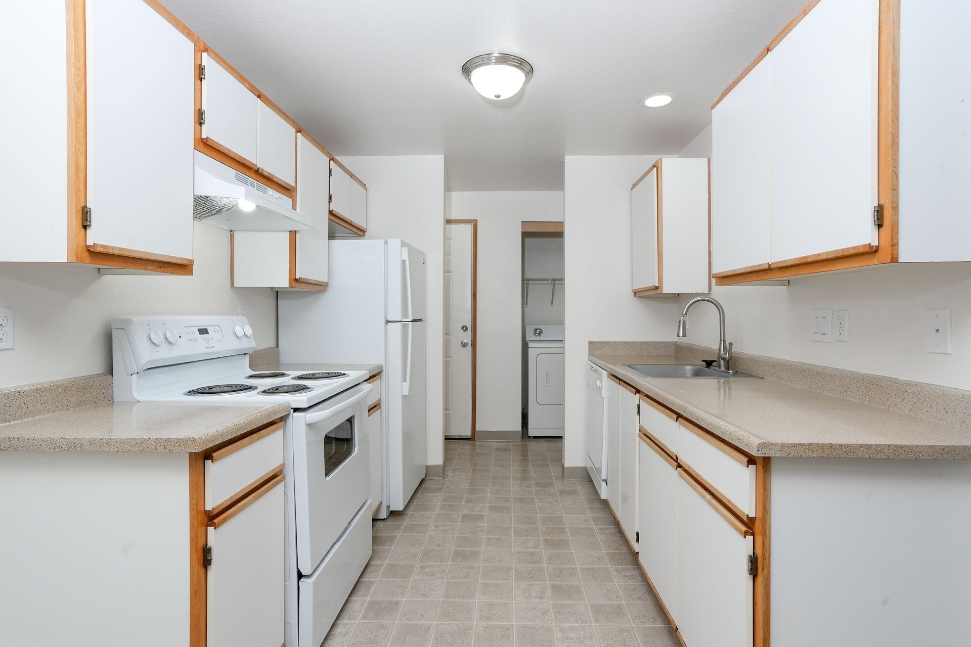 Bright white galley kitchen with white cabinets, beige countertops, stove, sink, and tiled floor.