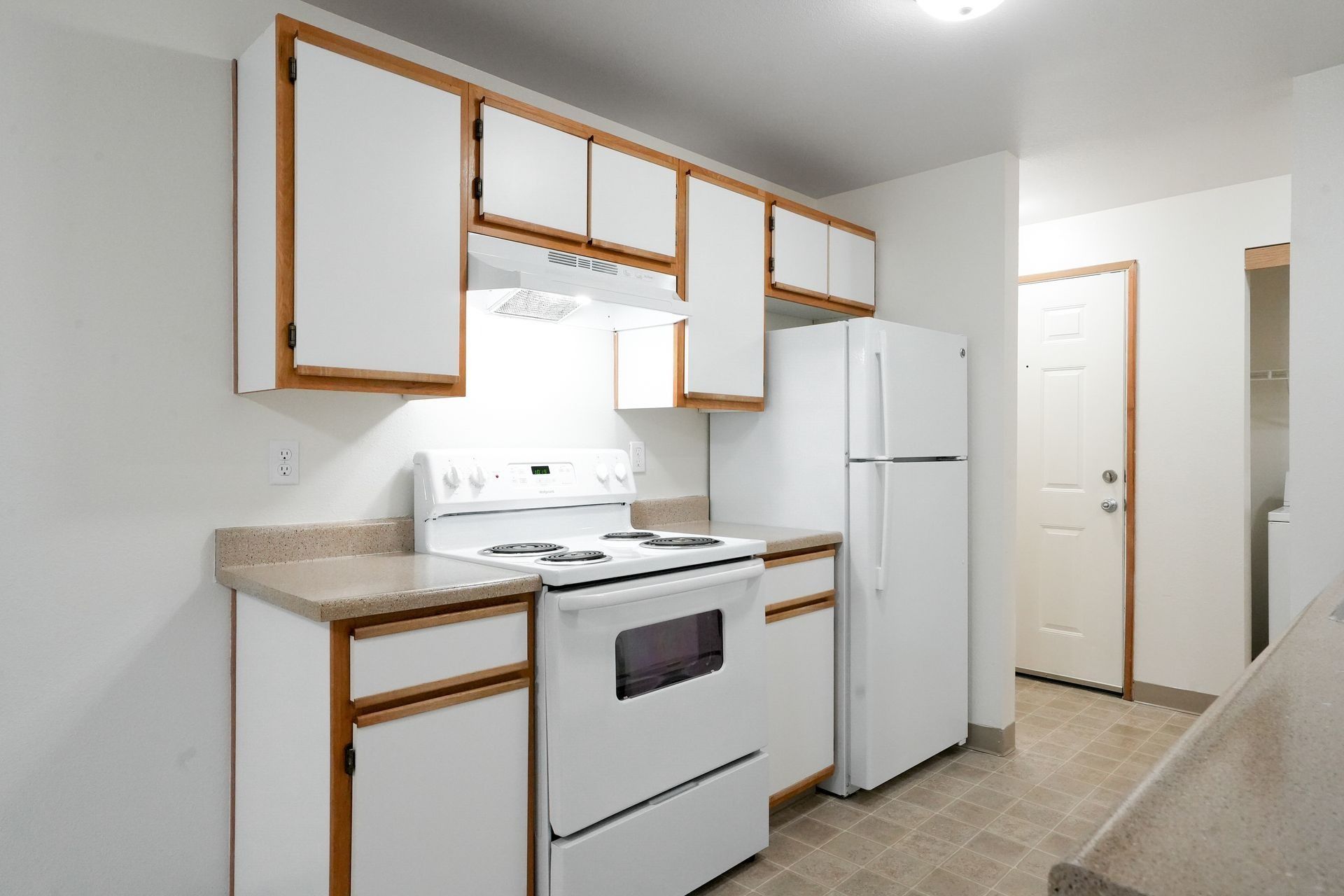 Small white kitchen with stove, fridge, and white cabinets along a narrow wall.