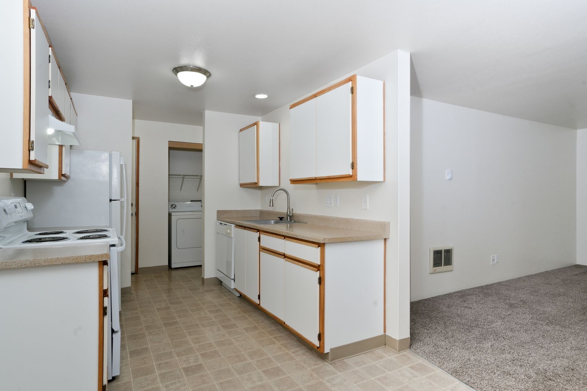 Empty kitchen and living area with white cabinets, tan counters, and beige carpet.