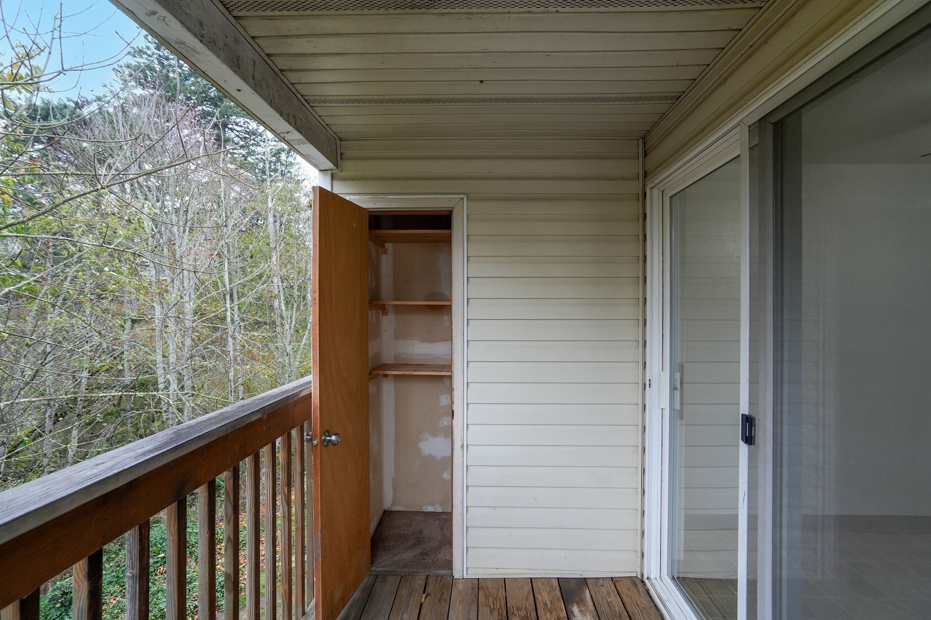 Covered wooden balcony with railing, sliding glass door, and open utility closet cabinet.