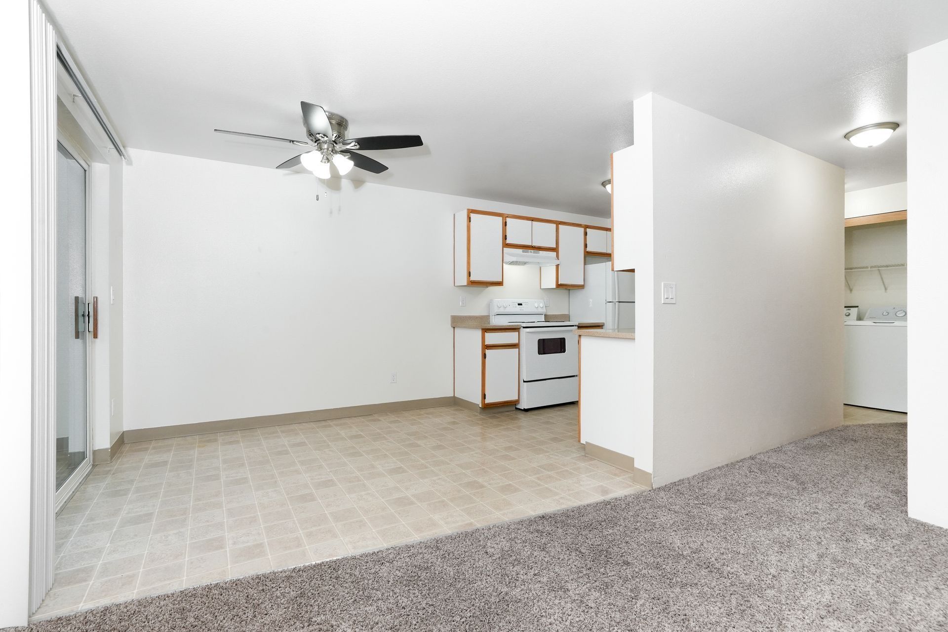 Empty apartment living room and kitchen with white walls, ceiling fan, and carpeted floor