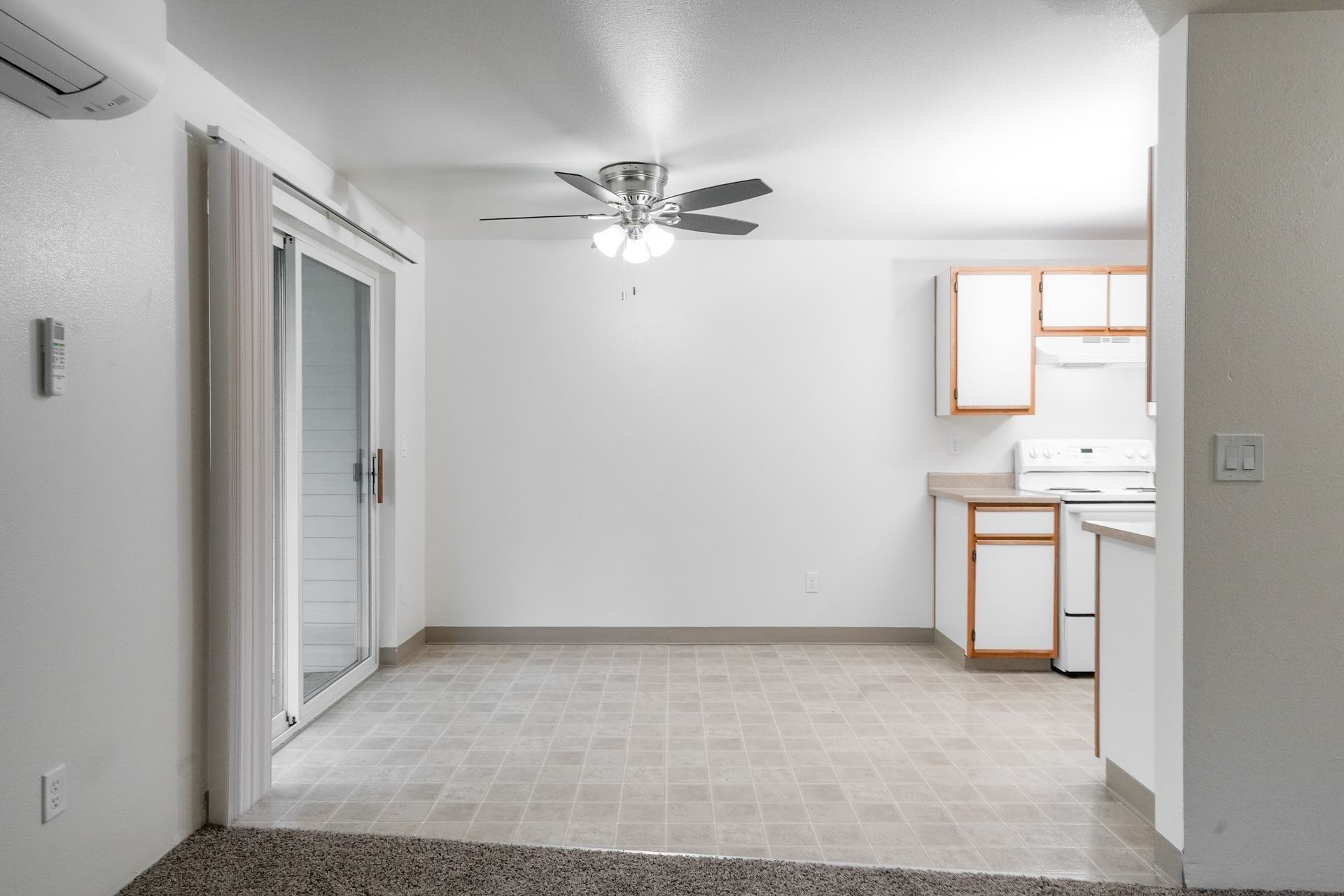 Empty living room with white walls, tiled floor, ceiling fan, and a small kitchen area on the right.