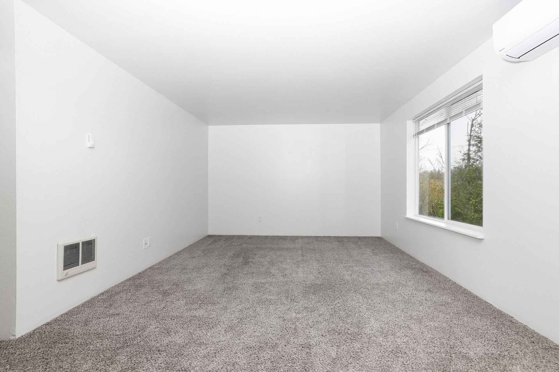 Empty white carpeted room with a window on the right and a wall vent on the left.