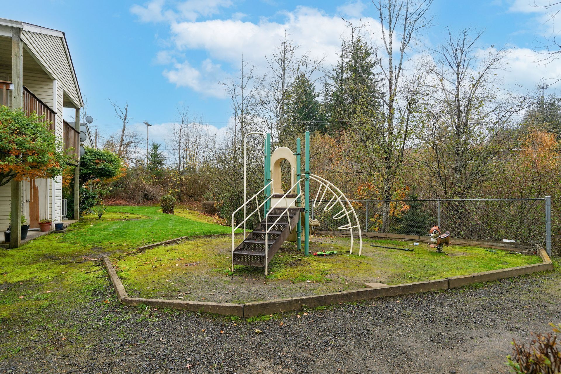 Backyard playground with a small slide, climbing frame, and fence, beside a house under a cloudy sky