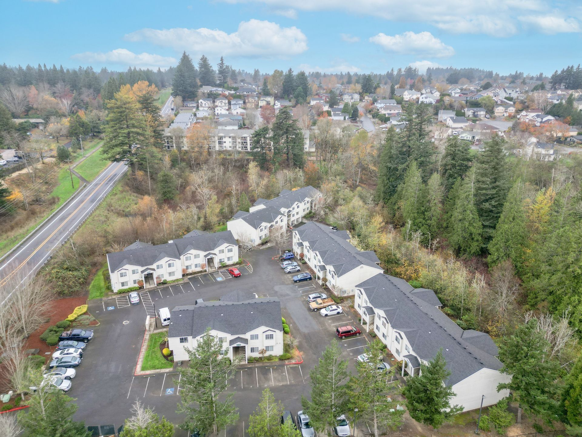 Aerial view of a suburban apartment complex beside a road and wooded area under a partly cloudy sky