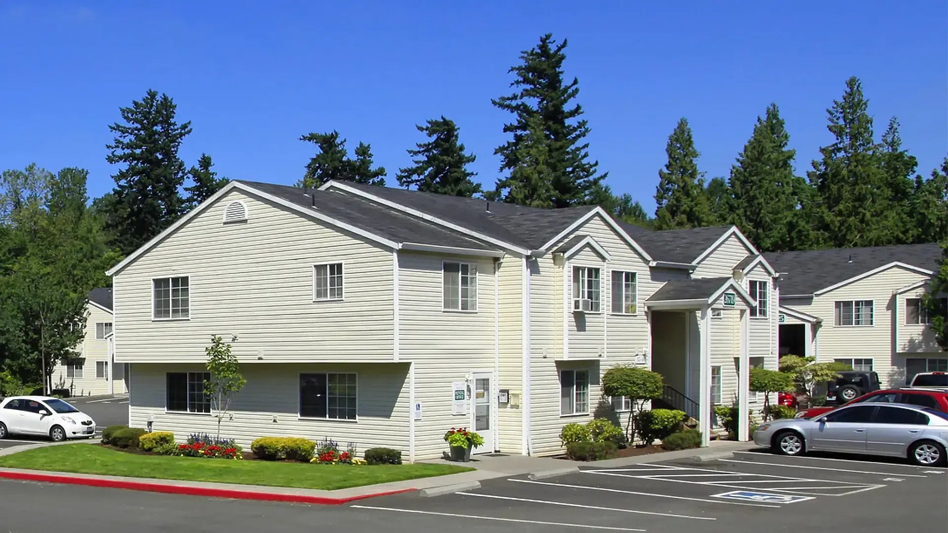 Apartment complex with beige siding, parked cars, and tall trees under a clear blue sky