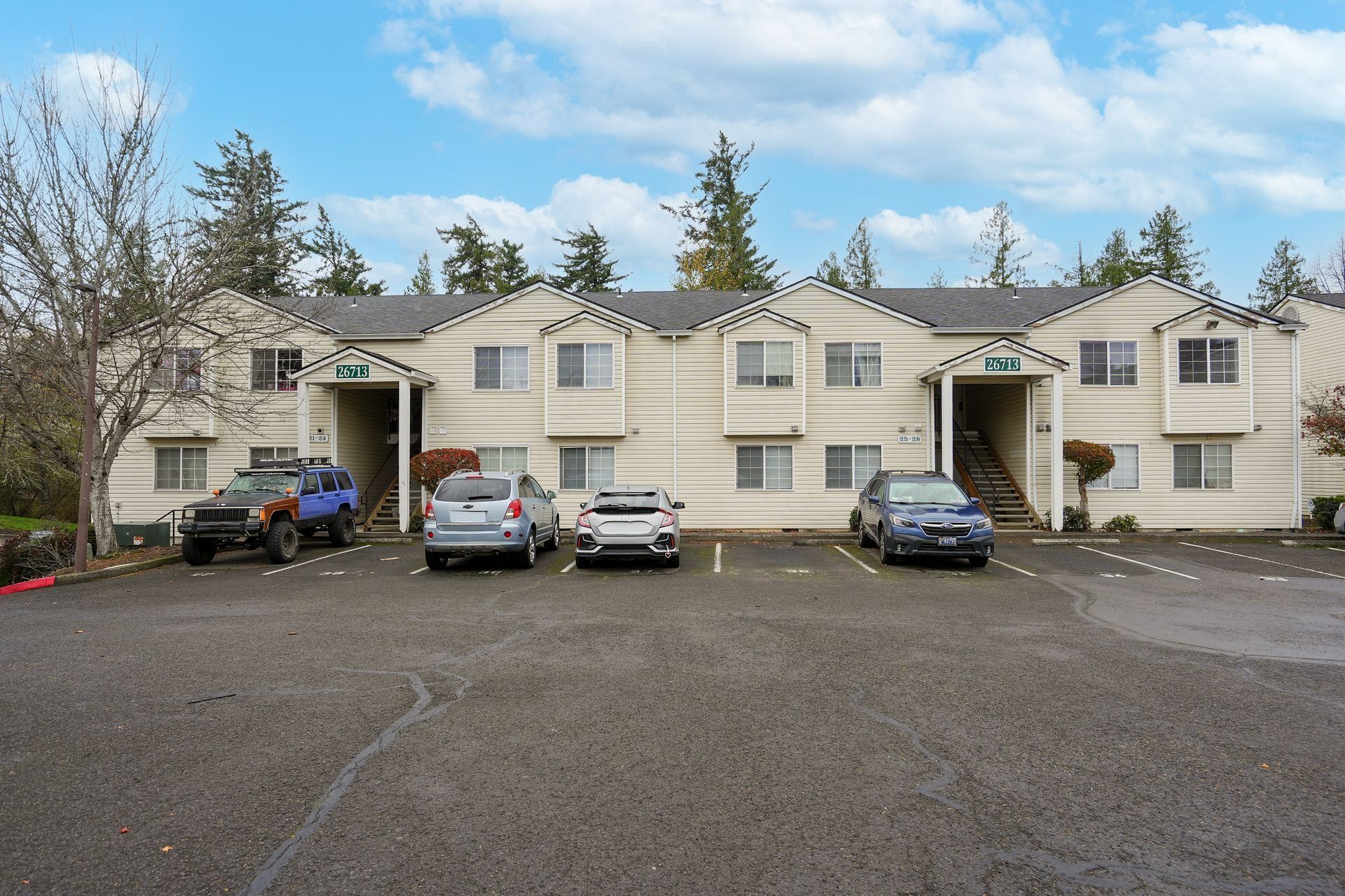Row of beige apartment buildings with parked cars in a gravel lot on a cloudy day