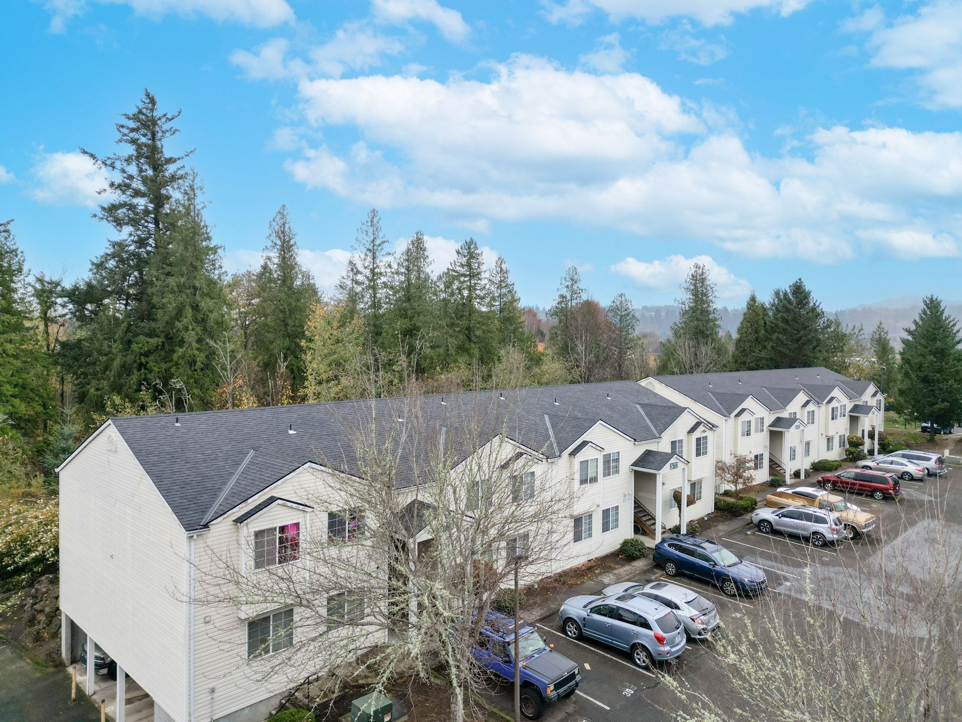 Aerial view of a white apartment building with parked cars and trees under a blue sky