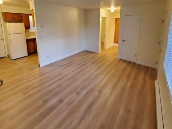 Empty apartment living room with wood floors, kitchen at left, and hallway to the right