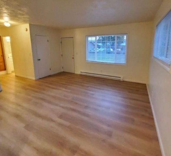 Empty living room with wood floors, beige walls, large window, and a hallway entrance.