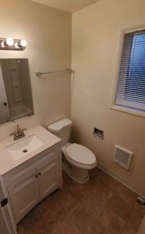 Small bathroom with white vanity, toilet, wall mirror, window blinds, and wood-look tile floor.