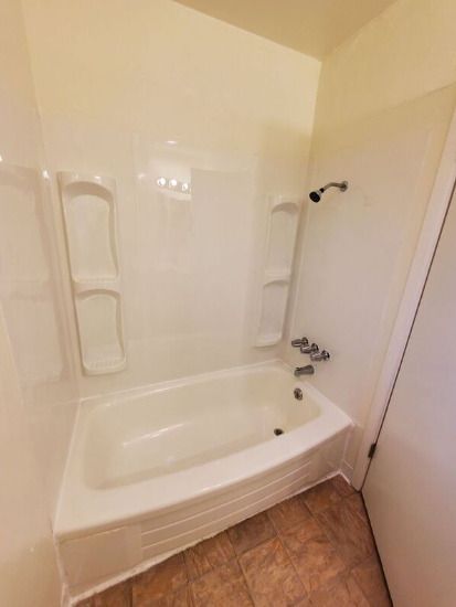 Small white bathtub with built-in grab bars and brown tile floor in a narrow bathroom