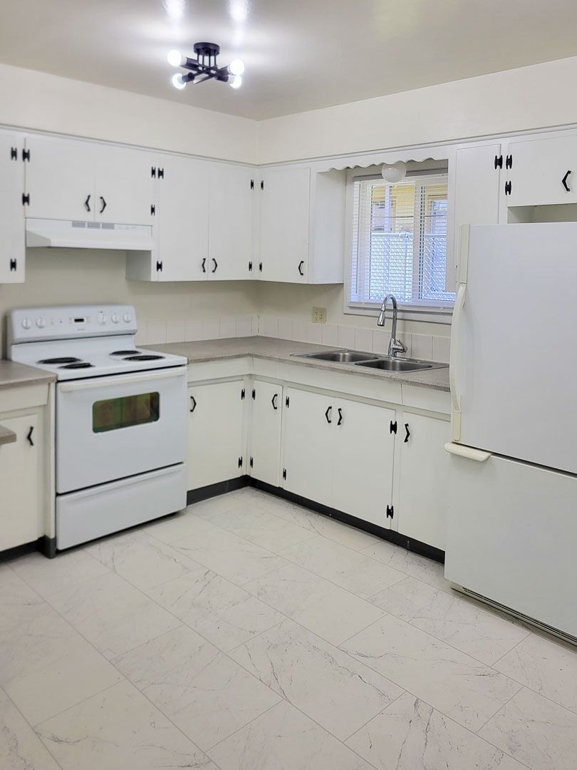 Bright white kitchen with stove, cabinets, fridge, and window over the sink
