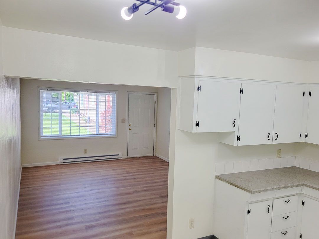 Empty room with wood floors, white cabinets, and a windowed alcove opening to a kitchen area