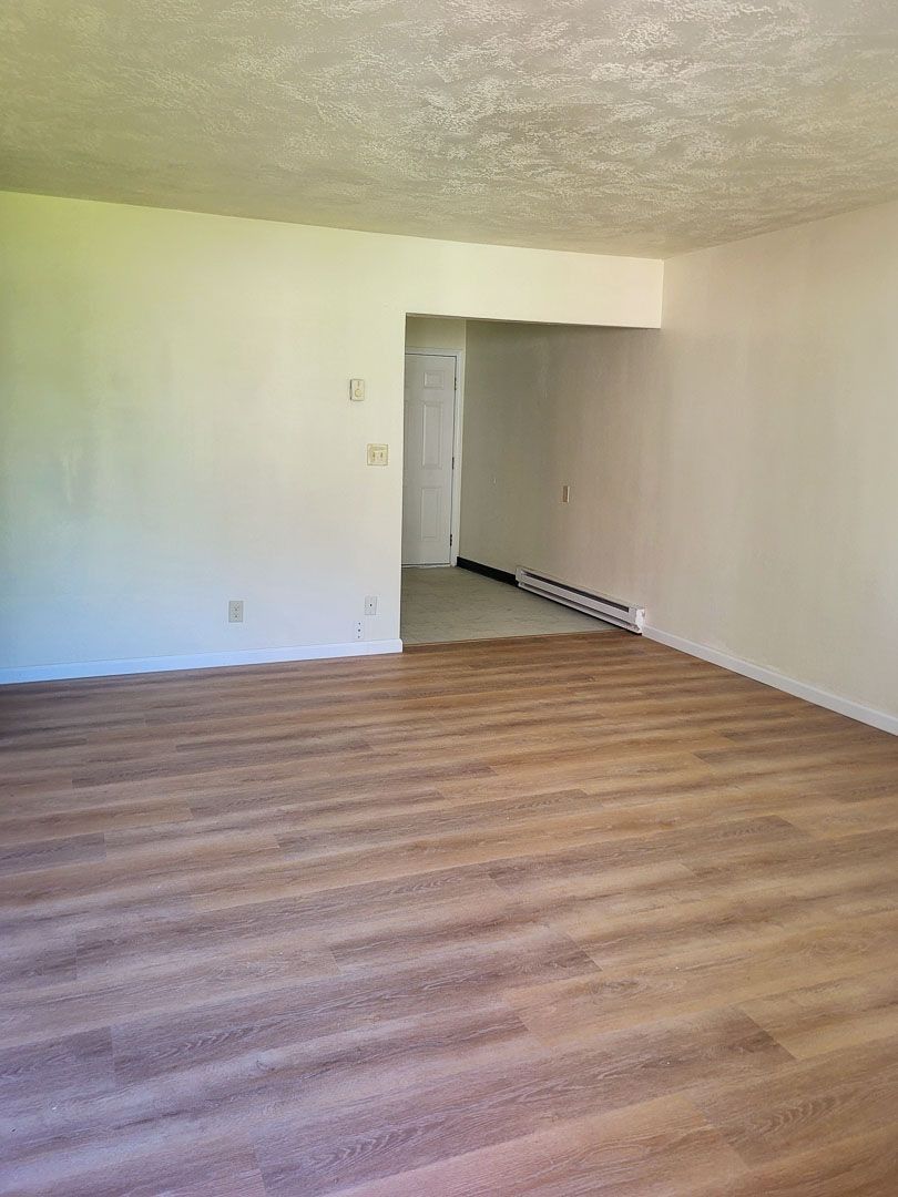 Empty room with striped wood floor, white walls, and a doorway to a hallway
